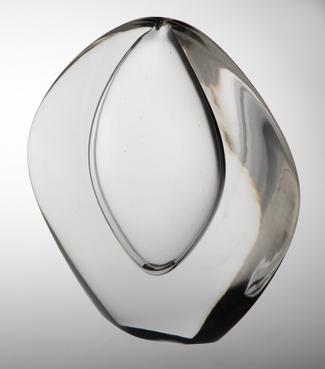 Vase av klart glass med skulpturell utforming. Flattrykket oval hovedform med konvekse sider. Ellipseformet hulerom i midten med en liten åpning på toppen.
