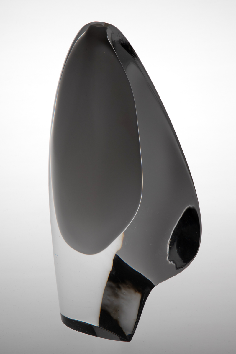 Skiveformet vase med en abstrakt, skulpturell utforming i underfangsglass. Det innerste sjiktet består av opakt melkehvitt glass som danner et tilnærmet elipseformet hulerom, mens det ytre sjiktet er i klart glass.