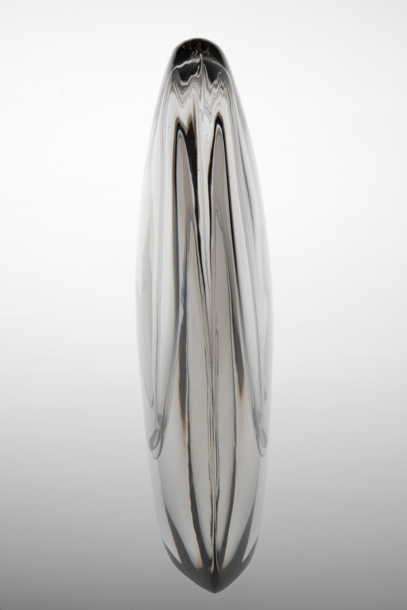 Vase av klart glass med skulpturell utforming. Flattrykket oval hovedform med konvekse sider. Ellipseformet hulerom i midten med en liten åpning på toppen.