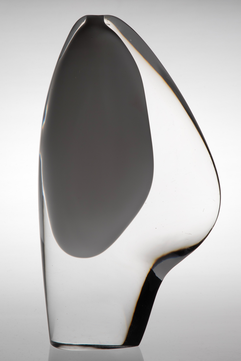 Skiveformet vase med en abstrakt, skulpturell utforming i underfangsglass. Det innerste sjiktet består av opakt melkehvitt glass som danner et tilnærmet elipseformet hulerom, mens det ytre sjiktet er i klart glass.