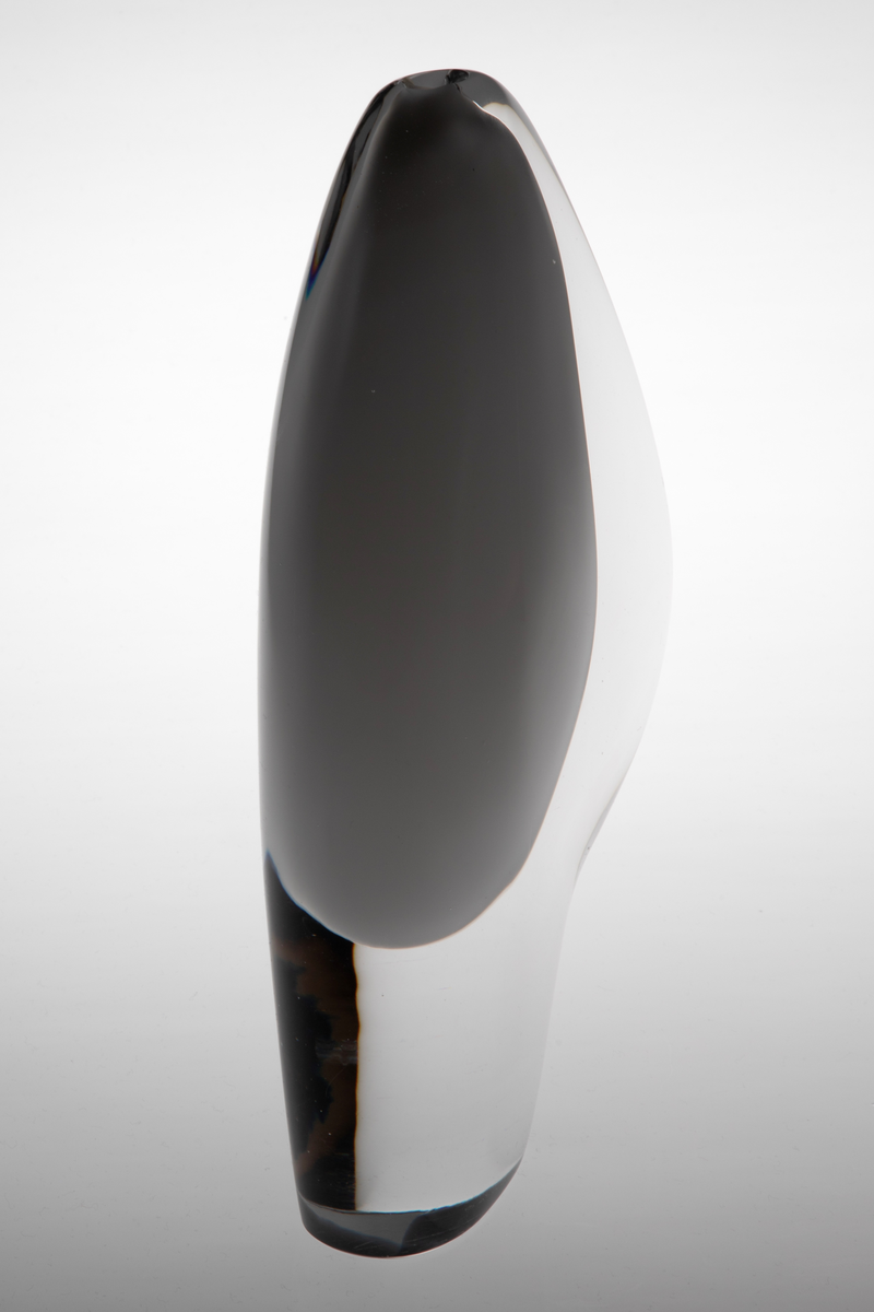 Skiveformet vase med en abstrakt, skulpturell utforming i underfangsglass. Det innerste sjiktet består av opakt melkehvitt glass som danner et tilnærmet elipseformet hulerom, mens det ytre sjiktet er i klart glass.