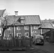 Motiv från Hunnebergsgatan 25 i Linköping 1961. Boningshuset med bod och uthus sedda från gården. Bilen är en Triumf Mayflower från 1951.