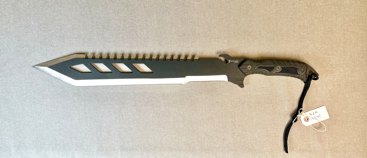 Tvegget stor kniv, med sort matt perforert blad som avsluttes i tveegget spiss. Den øvre eggen har vinklede tagger (mothaker). Sort gummiert håndtak. Slire i tykt sort nylonstoff, som lukkes med trykknapper. Slren er forterket med aluminium i spissen.