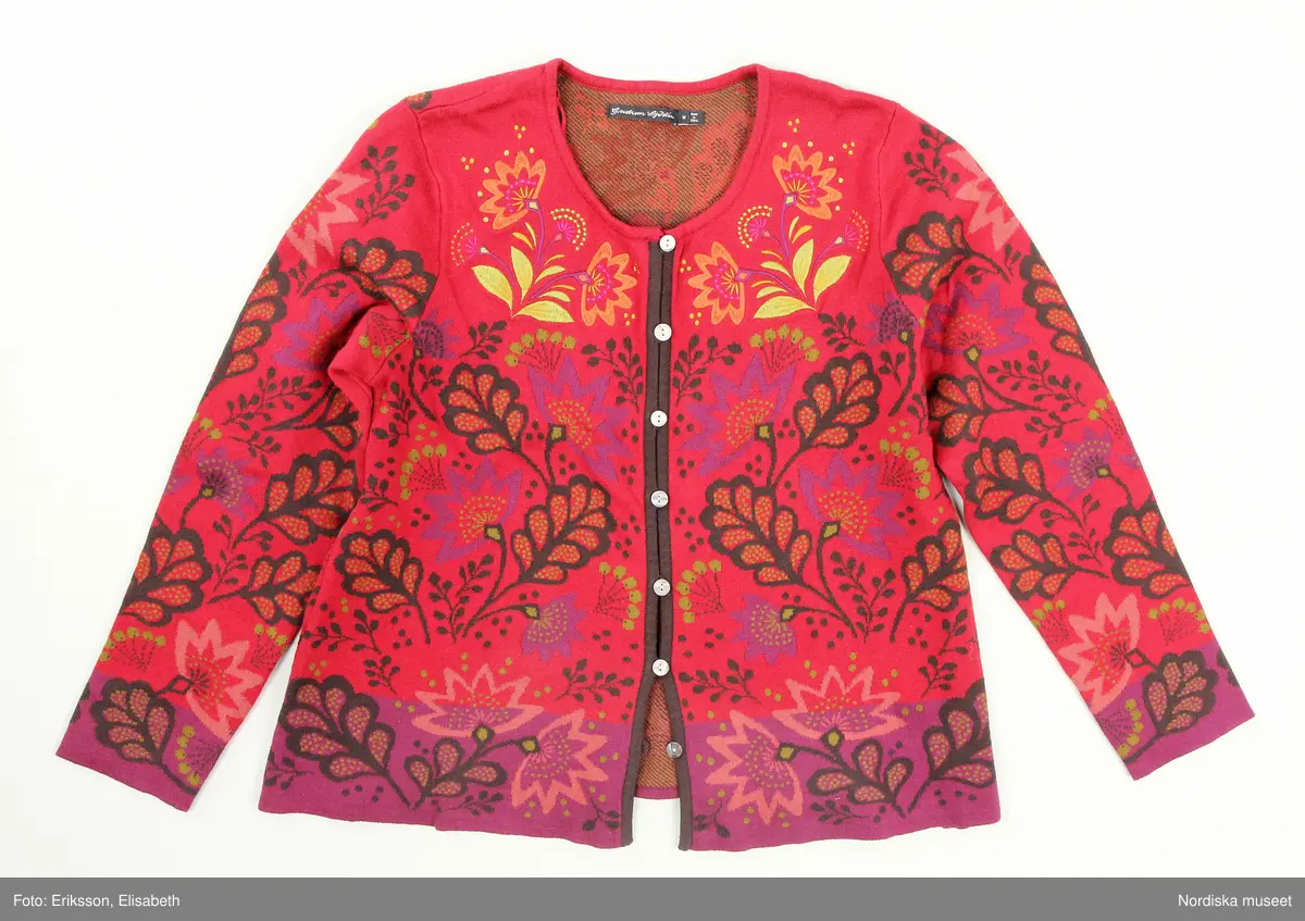 Kofta, stickad (trikå) av ekologisk bomull. Rak modell. Röd botten med stiliserade vegetativa mönster i färger. I nederkant och i ärmslut lila botten. Runt halslinningen broderade blommor. Bandkantad knäppning fram med sju knappar av pärlemor och tränsar. Ett par hankar på axelsömmarnas insida (trol. för att användas att fästa på galge i affär, så koftan inte ramlar av vid hantering). 
I nacken svarta tygetiketter med text: "Gudrun Sjödén  / M (=storleksangivelse för medium), Made in China".
I vänster sidsöm vit etikett med med materialdeklaration, tvättinstruktioner och uppgift om att plagget innehåller "icketextila delar av animaliskt ursprung". På etiketten fäst extraknapp.
/Leif Wallin 2025-02-19