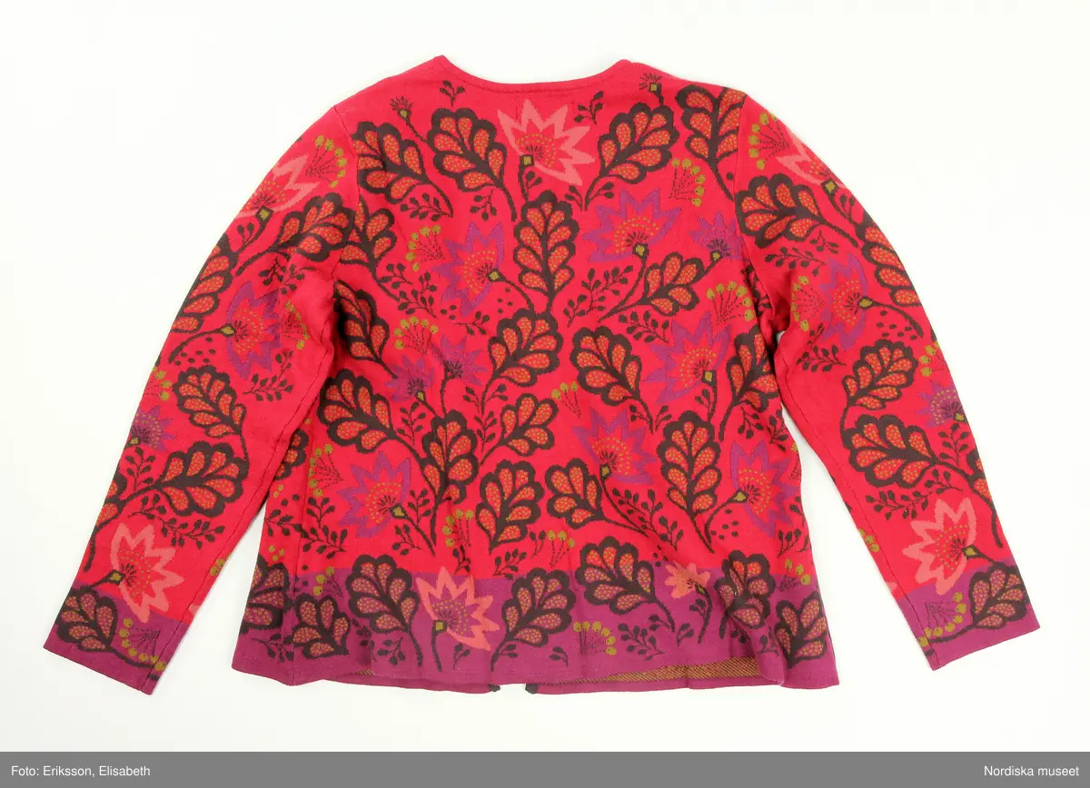 Kofta, stickad (trikå) av ekologisk bomull. Rak modell. Röd botten med stiliserade vegetativa mönster i färger. I nederkant och i ärmslut lila botten. Runt halslinningen broderade blommor. Bandkantad knäppning fram med sju knappar av pärlemor och tränsar. Ett par hankar på axelsömmarnas insida (trol. för att användas att fästa på galge i affär, så koftan inte ramlar av vid hantering). 
I nacken svarta tygetiketter med text: "Gudrun Sjödén  / M (=storleksangivelse för medium), Made in China".
I vänster sidsöm vit etikett med med materialdeklaration, tvättinstruktioner och uppgift om att plagget innehåller "icketextila delar av animaliskt ursprung". På etiketten fäst extraknapp.
/Leif Wallin 2025-02-19