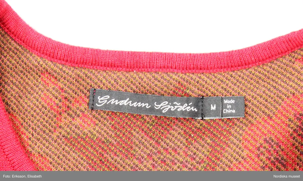 Kofta, stickad (trikå) av ekologisk bomull. Rak modell. Röd botten med stiliserade vegetativa mönster i färger. I nederkant och i ärmslut lila botten. Runt halslinningen broderade blommor. Bandkantad knäppning fram med sju knappar av pärlemor och tränsar. Ett par hankar på axelsömmarnas insida (trol. för att användas att fästa på galge i affär, så koftan inte ramlar av vid hantering). 
I nacken svarta tygetiketter med text: "Gudrun Sjödén  / M (=storleksangivelse för medium), Made in China".
I vänster sidsöm vit etikett med med materialdeklaration, tvättinstruktioner och uppgift om att plagget innehåller "icketextila delar av animaliskt ursprung". På etiketten fäst extraknapp.
/Leif Wallin 2025-02-19