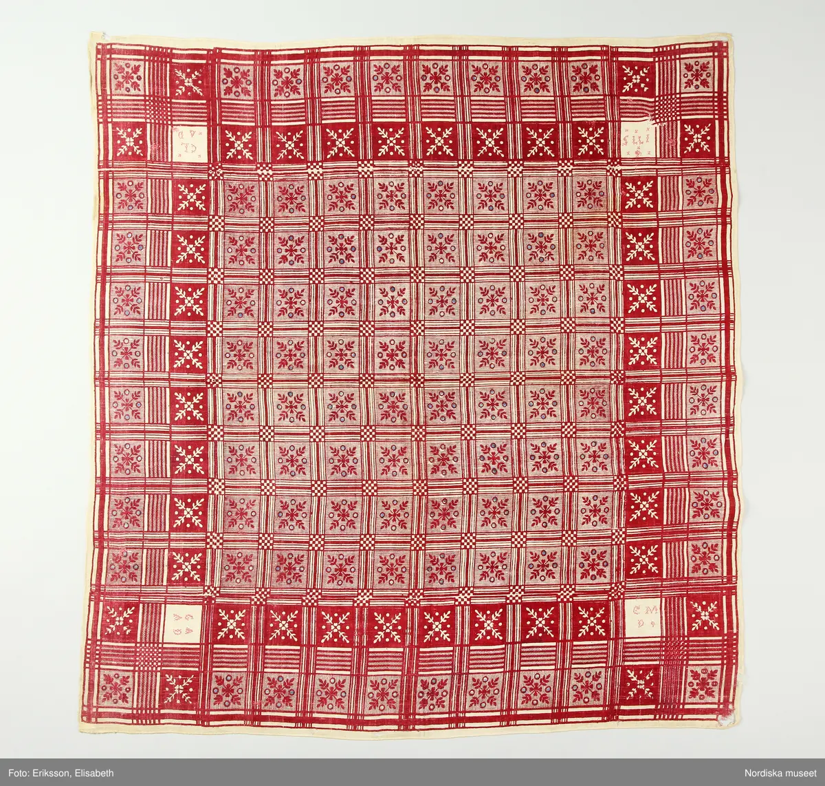 Halskläde av bomull, 79 x 84 cm, vävt i tunn, gles tuskaft. Blocktryckt mönster tryckt med rött mot oblekt botten, med glesa inslag av indigoblått. Mönstret indelat i rutor med stiliserade växtornament. I varje hörn en sparad oblekt ruta med broderi i rosa korssöm, en ruta visar årtalet 1775, övriga tre rutor initialerna G A A D, C M D respektive C L A D (något otydligt). Längs sidorna en smal fåll, fållad för hand med vit tråd.
Anm. Sliten och trasig, framför allt i hörnen, vissa hål över ytan lagade med röd respektive vit tråd till synes under olika tidsskeden.
/Marianne Larsson 2024-09-30