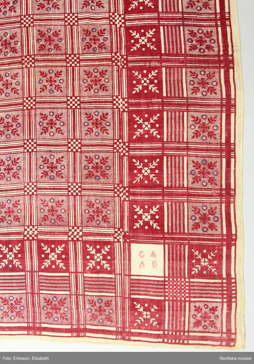 Halskläde av bomull, 79 x 84 cm, vävt i tunn, gles tuskaft. Blocktryckt mönster tryckt med rött mot oblekt botten, med glesa inslag av indigoblått. Mönstret indelat i rutor med stiliserade växtornament. I varje hörn en sparad oblekt ruta med broderi i rosa korssöm, en ruta visar årtalet 1775, övriga tre rutor initialerna G A A D, C M D respektive C L A D (något otydligt). Längs sidorna en smal fåll, fållad för hand med vit tråd.
Anm. Sliten och trasig, framför allt i hörnen, vissa hål över ytan lagade med röd respektive vit tråd till synes under olika tidsskeden.
/Marianne Larsson 2024-09-30