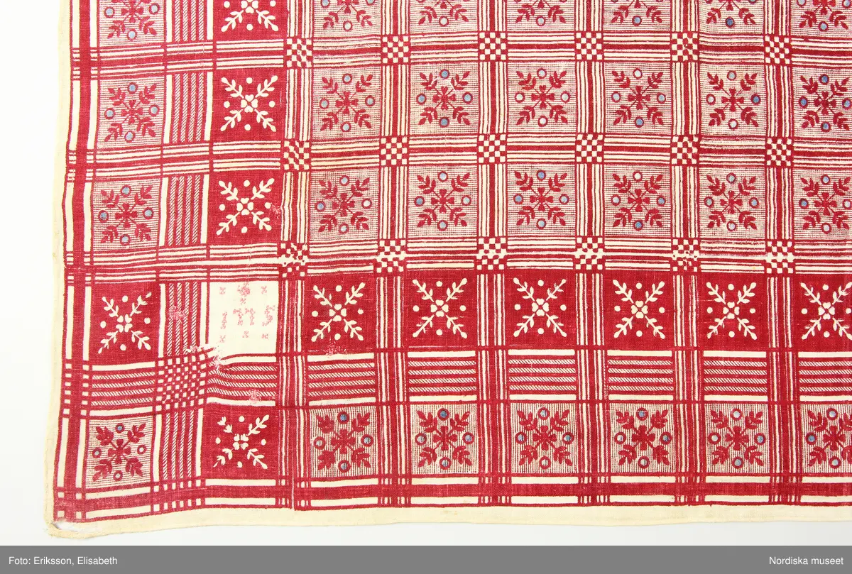 Halskläde av bomull, 79 x 84 cm, vävt i tunn, gles tuskaft. Blocktryckt mönster tryckt med rött mot oblekt botten, med glesa inslag av indigoblått. Mönstret indelat i rutor med stiliserade växtornament. I varje hörn en sparad oblekt ruta med broderi i rosa korssöm, en ruta visar årtalet 1775, övriga tre rutor initialerna G A A D, C M D respektive C L A D (något otydligt). Längs sidorna en smal fåll, fållad för hand med vit tråd.
Anm. Sliten och trasig, framför allt i hörnen, vissa hål över ytan lagade med röd respektive vit tråd till synes under olika tidsskeden.
/Marianne Larsson 2024-09-30