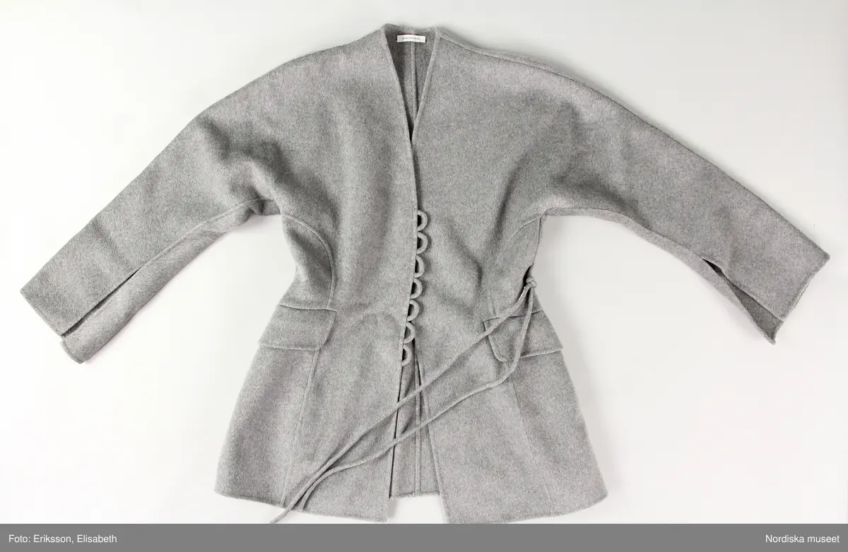 Jacka av filtad grå ylle med lös snodd/skärp (a-b). Två framstycken, två sidstycken och två bakstycken. Ärm med tre sömmar. Ärmen skuren i ett stycke med fram respektive bakstycke. Ärmen med slits nedtill. 
Jackan något insvängd i midjan. En ficka med ficklock på var framtstycke. På vänster framstyckes framkant sju stycken öglor och på höger framstycke en ögla vari fäst lös snodd (b) av samma material för stängning. 
Ofodrad och kraglös. 
På insidan fickpåsar av ull samt satin. 
Etikett i nacken med text "BY MALENE BIRGER" och liten etikett i vänster sidsöm ned storleksangivelse "36"
Fickorna ej sprättade.
/Leif Wallin 2024-12-11