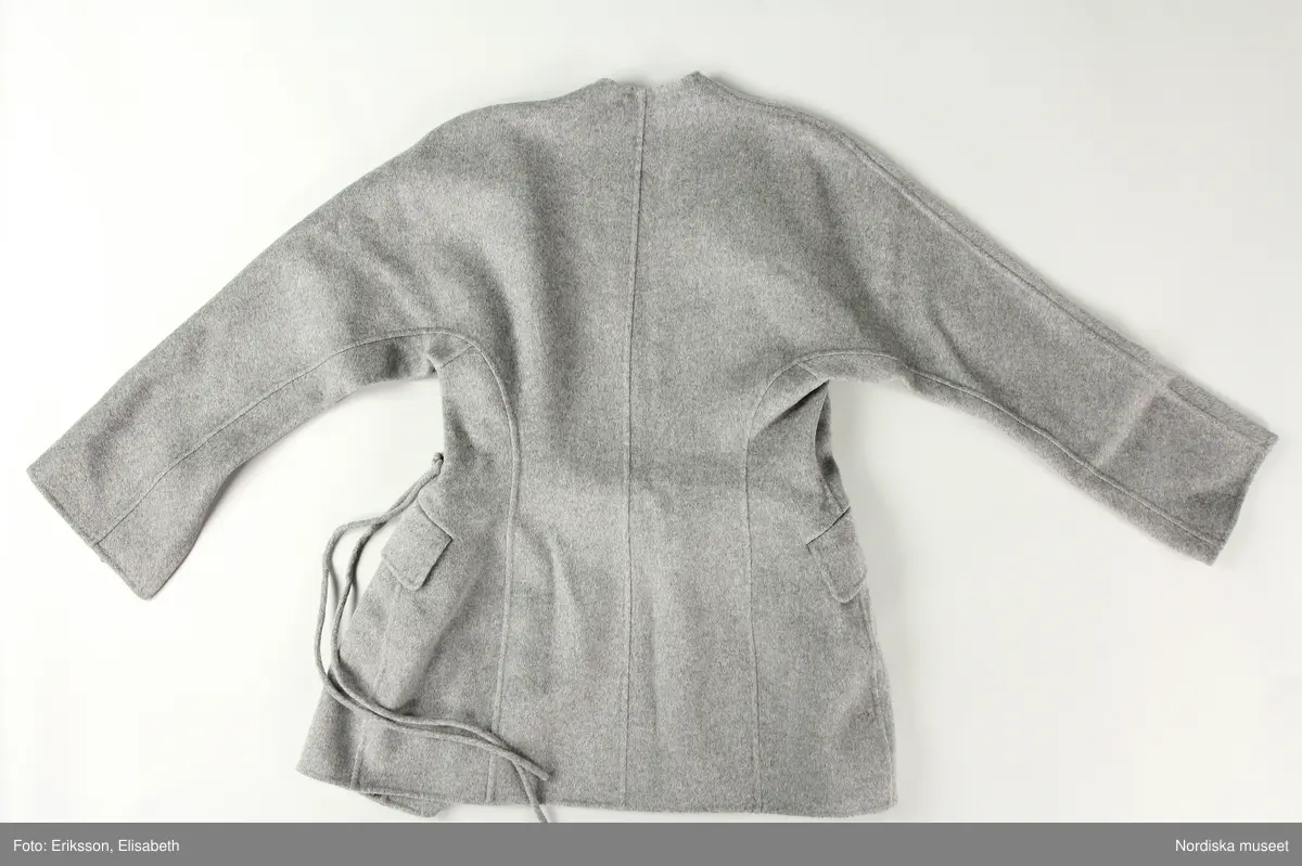 Jacka av filtad grå ylle med lös snodd/skärp (a-b). Två framstycken, två sidstycken och två bakstycken. Ärm med tre sömmar. Ärmen skuren i ett stycke med fram respektive bakstycke. Ärmen med slits nedtill. 
Jackan något insvängd i midjan. En ficka med ficklock på var framtstycke. På vänster framstyckes framkant sju stycken öglor och på höger framstycke en ögla vari fäst lös snodd (b) av samma material för stängning. 
Ofodrad och kraglös. 
På insidan fickpåsar av ull samt satin. 
Etikett i nacken med text "BY MALENE BIRGER" och liten etikett i vänster sidsöm ned storleksangivelse "36"
Fickorna ej sprättade.
/Leif Wallin 2024-12-11