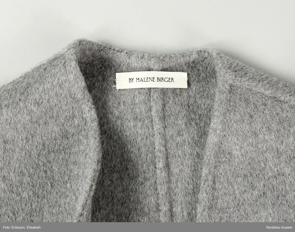 Jacka av filtad grå ylle med lös snodd/skärp (a-b). Två framstycken, två sidstycken och två bakstycken. Ärm med tre sömmar. Ärmen skuren i ett stycke med fram respektive bakstycke. Ärmen med slits nedtill. 
Jackan något insvängd i midjan. En ficka med ficklock på var framtstycke. På vänster framstyckes framkant sju stycken öglor och på höger framstycke en ögla vari fäst lös snodd (b) av samma material för stängning. 
Ofodrad och kraglös. 
På insidan fickpåsar av ull samt satin. 
Etikett i nacken med text "BY MALENE BIRGER" och liten etikett i vänster sidsöm ned storleksangivelse "36"
Fickorna ej sprättade.
/Leif Wallin 2024-12-11