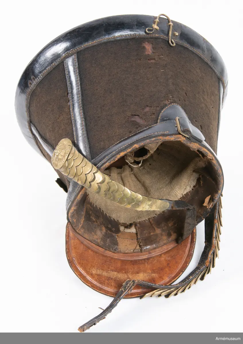 Ry. kiver. Av svart kläde och svart lackerat läder. Övre delen har rund, rak botten. Skillnaden mellan diametrarna på övre och nedre delen är 11.1 cm. På framsidan finns hållare för ståndare. Vapenplåt på tschakåns främre del av mässing; med ingraverad dubbelörn med en S:t Göransbild i mitten. Skärm av lackerat läder. Hakrem av mässingsplåtar. En knapp på hakremmen är av en annan modell än de andra. Foder av grovt brunt läder.