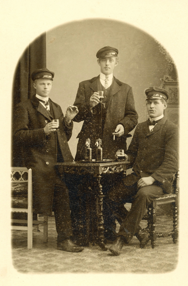 Ateljéfoto med tre unga män i vegamössor poserar med ölglas och cigaretter. På bordet står ett par buteljer. Uppgifter saknas.