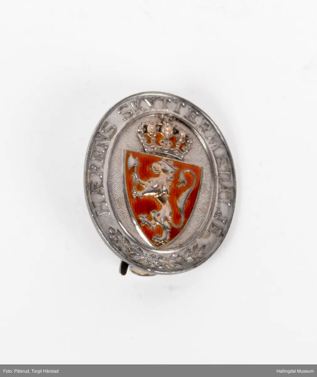 Hærens skyttermedalje. En nål med riksløven på emaljert rød bakgrunn, med skrift rundt. Laget i sølv.