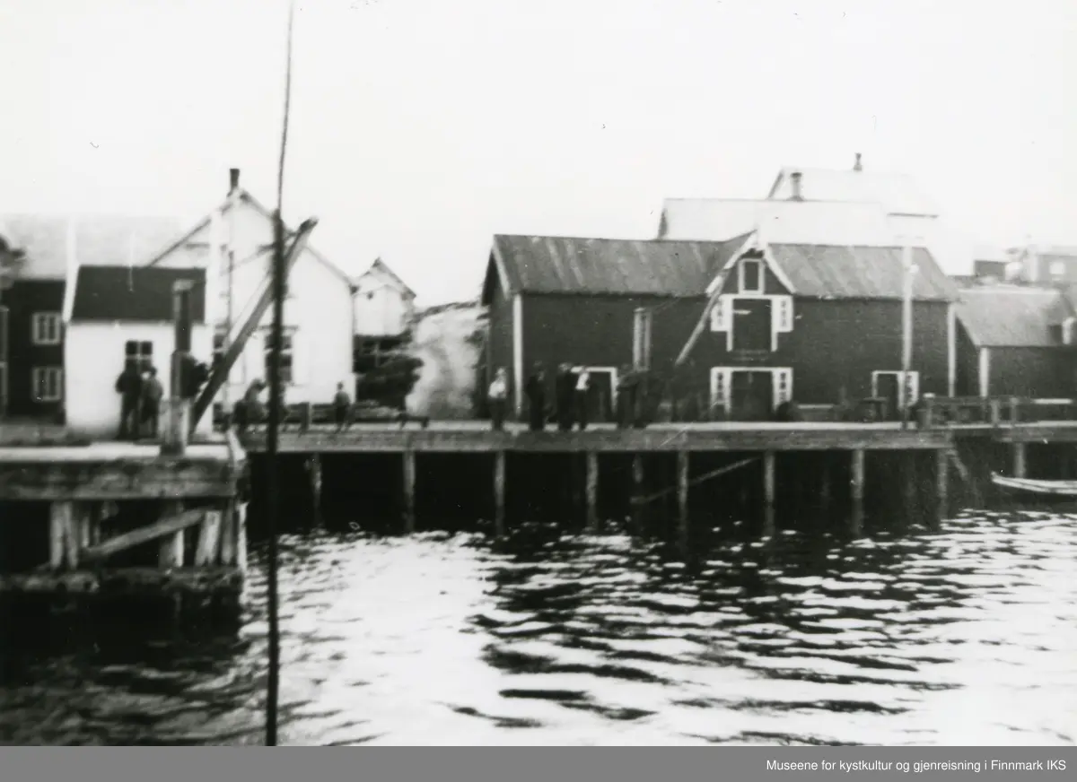 Gjesvær. Det gamle Nissenbruket og deler av bebyggelsen. Flere personer på kaia.