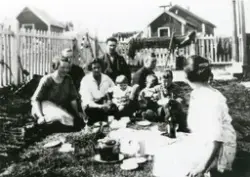 Flere voksne og barn har en piknik i en inngjerdet hage. I bakgrunnen deler av bebyggelsen. Til høyre ser man husinngangen. Antatt Gjesvær.