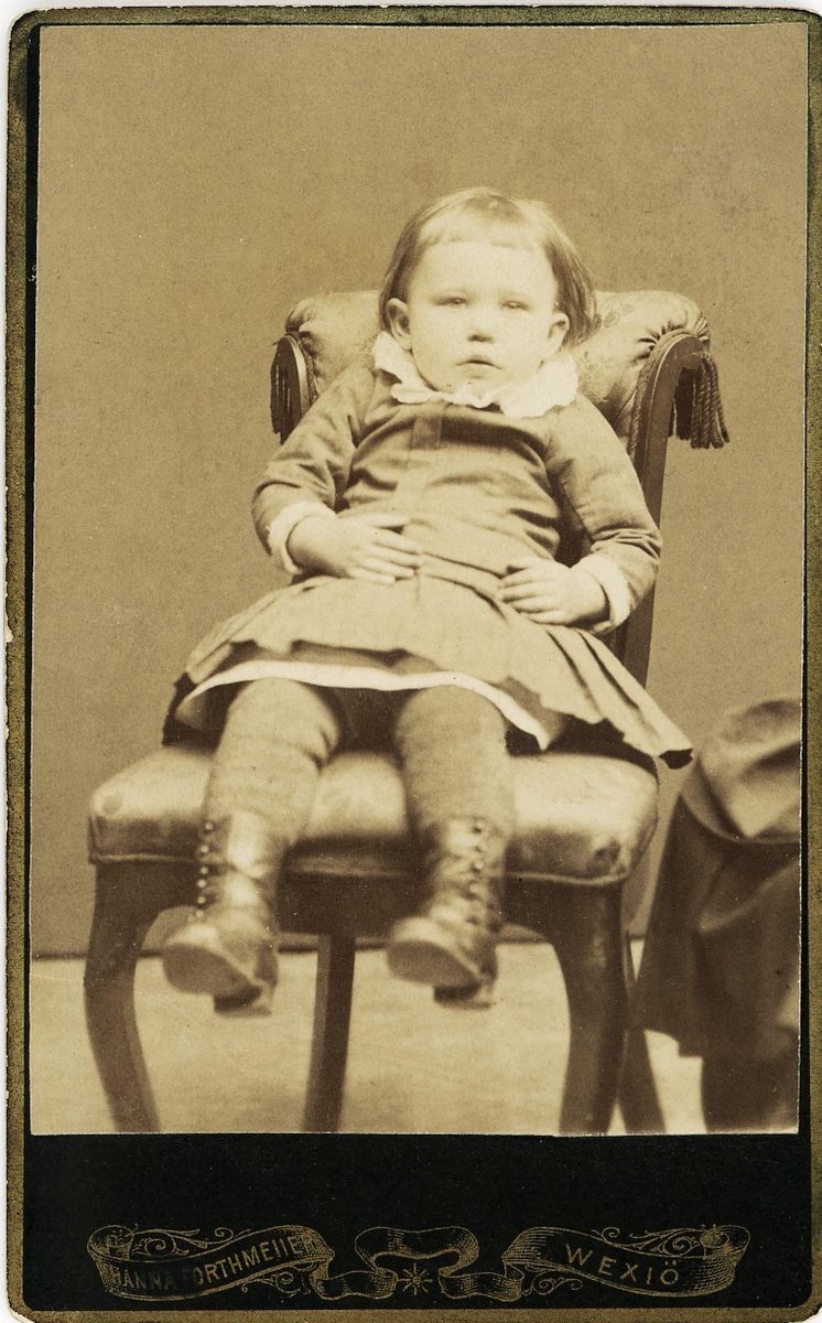 En liten pojke i kolt sitter på en stoppad stol hos fotografen, cirka 1875.
Helfigur, en face. Ateljéfoto.