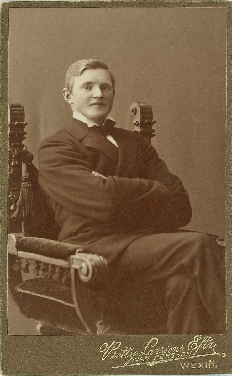 En man i mörk kostym med väst och stärkkrage med fluga, sitter i en utsirad karmstol hos fotografen.
Knäbild, halvprofil. Ateljéfoto.