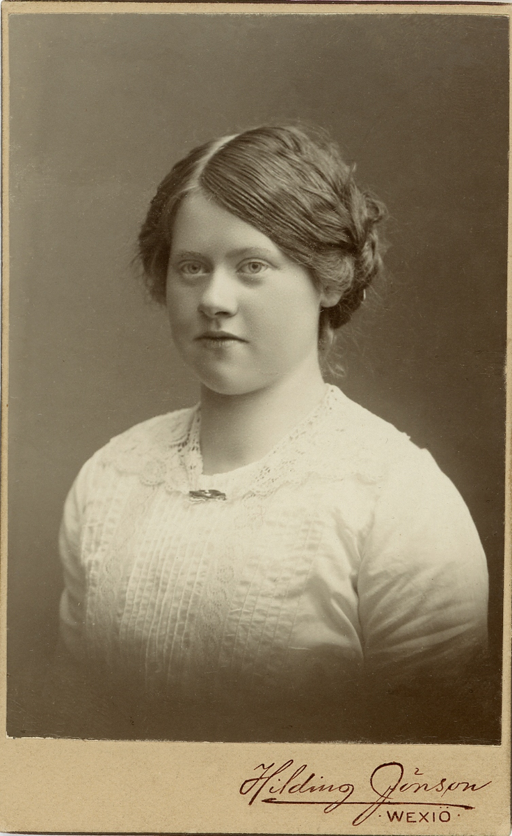 En ung kvinna i ljus blus med stråveck, spetskrage och en liten brosch. 
På baksidan står antecknat: "Dotter till EI" (?). 
Bröstbild, halvprofil. Ateljéfoto.