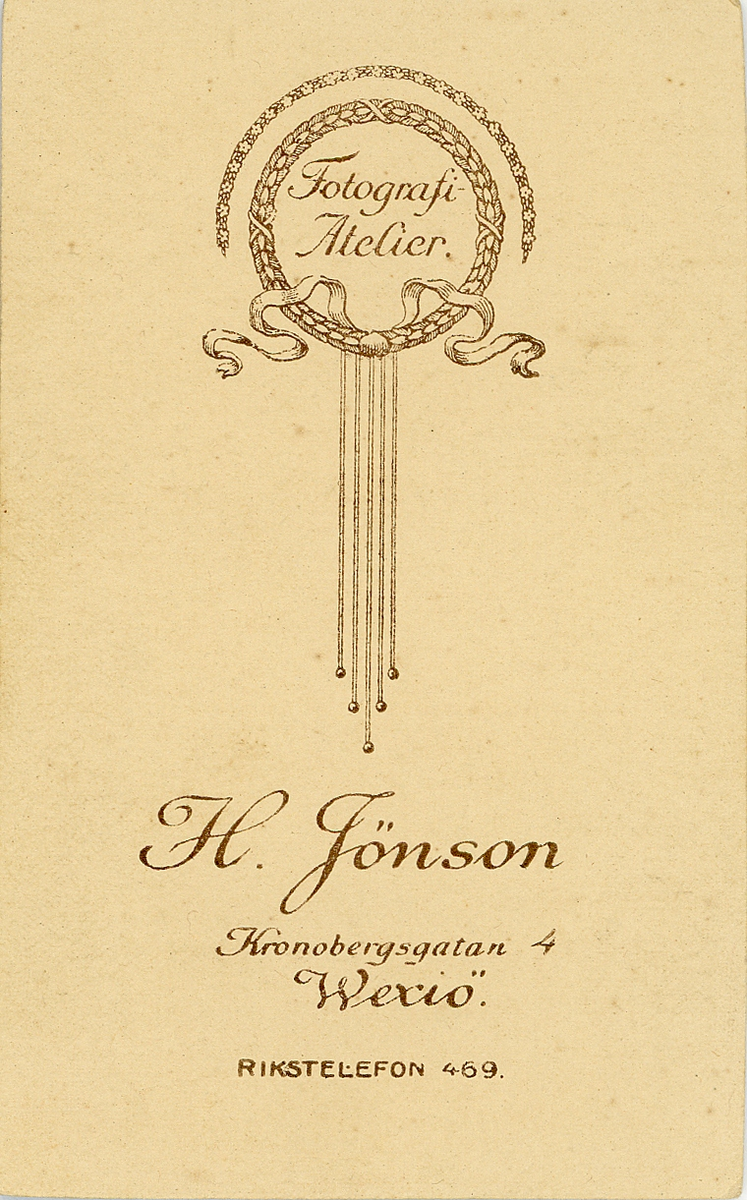 En ung kvinna i mörk, höghalsad klänning med intrikata broderier, halskedja och vid halsgropen syns en brosch med snirkliga initialer. 
Bröstbild, halvprofil. Ateljéfoto.