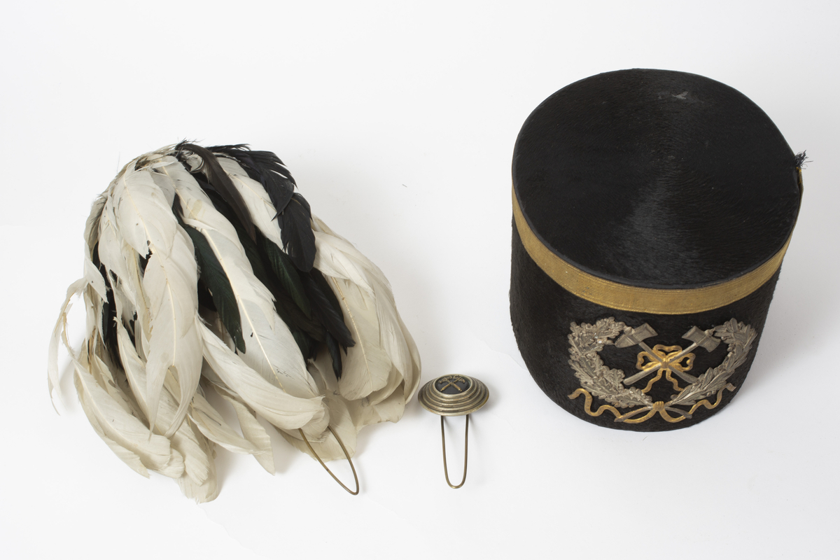 Tradisjonel tysk bergmannshatt, "Schachthut" til paradeuniform. Trukket med fløyel. Merke i metall med hammer og jern-symbolet og en krans med sløyfe foran. Gullfarget bånd rundt toppkanten. Fjærbusk med hvite og sorte fjær. Feste til denne er i metall og har en rund knapp med hammer og jern-symbolet. Hatten har et hull på venstre side hvor denne stikkes nedi. Foret med lyst silke(?)stoff. Svettebånd av brunt skinn.