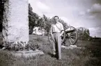 Foto av man vid en minnessten med militär kanon bakom sig. Text på stenen: Till minne av Vaxö läger reste vapenbröder och Vaxholms stad denna sten år 1939. Minnesstenen står i backen upp mot storstugan på Hamngatan i Vaxholm.