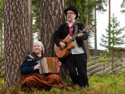 To musikere i historiske klær spiller trekkspill og gitar i skoglandskap.