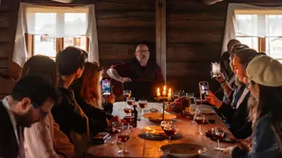 En mann sitter i enden av et langbord og synger til en gruppe som sitter ved langbordet. Det er mat, drikke og tente lys på bordet.