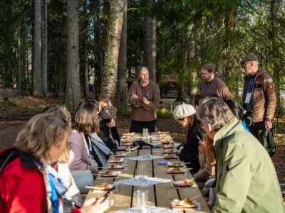 En gruppe voksne sitter på et langbord ute i naturen og følger med på en mann som står og snakker