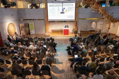 En person holder en presentasjon foran et større publikum i en sal. 