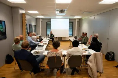 En gruppe voksne sitter i en hestesko og følger med på en presenasjon