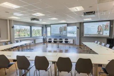 Et stort rom med bord og stoler satt i hesteskoformasjon foran to skjermer