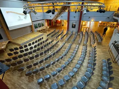 Et stort rom med mange stoler plassert i halvbuer eterhverandre foran en stor skjerm