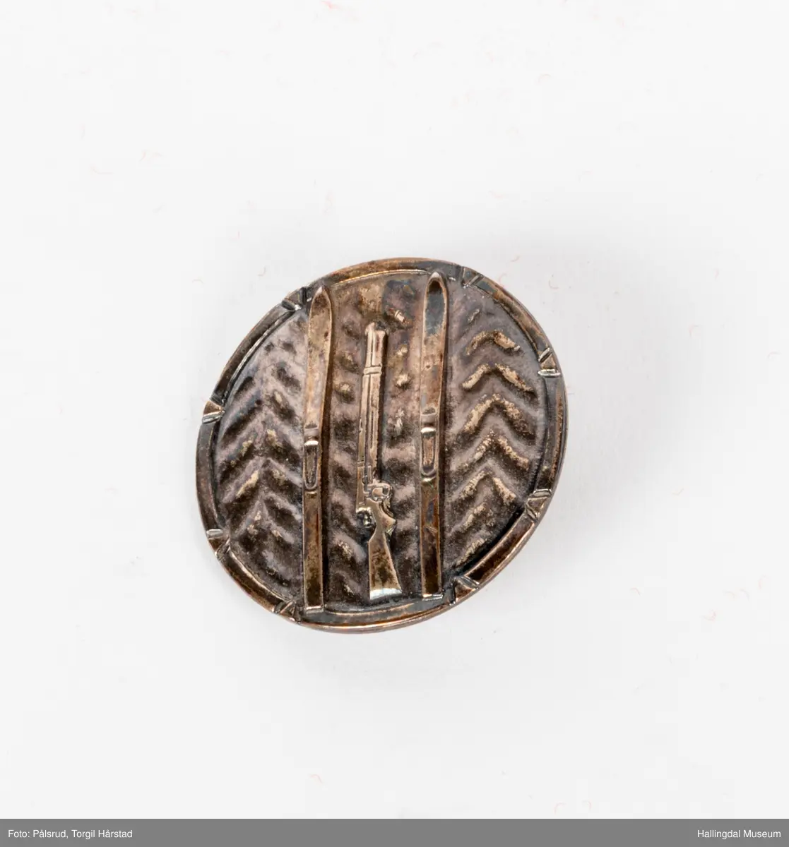 Det militære skimerke 1915-1945, en rund nål i sølv. Motivet er to ski og et gevær i mellom de, som står på en bakgrunn med det som skal forestille trær.