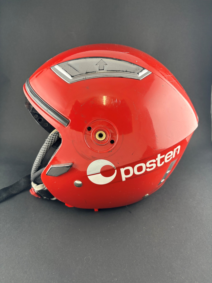Rød hjelm med tilhørende pose for oppbevaring. Merket med Postens logomerke. Hjelmen mangler visir.

Oppbevaringspose for hjelmen med postens logomerke. Rød og hvit med snor til lukking.