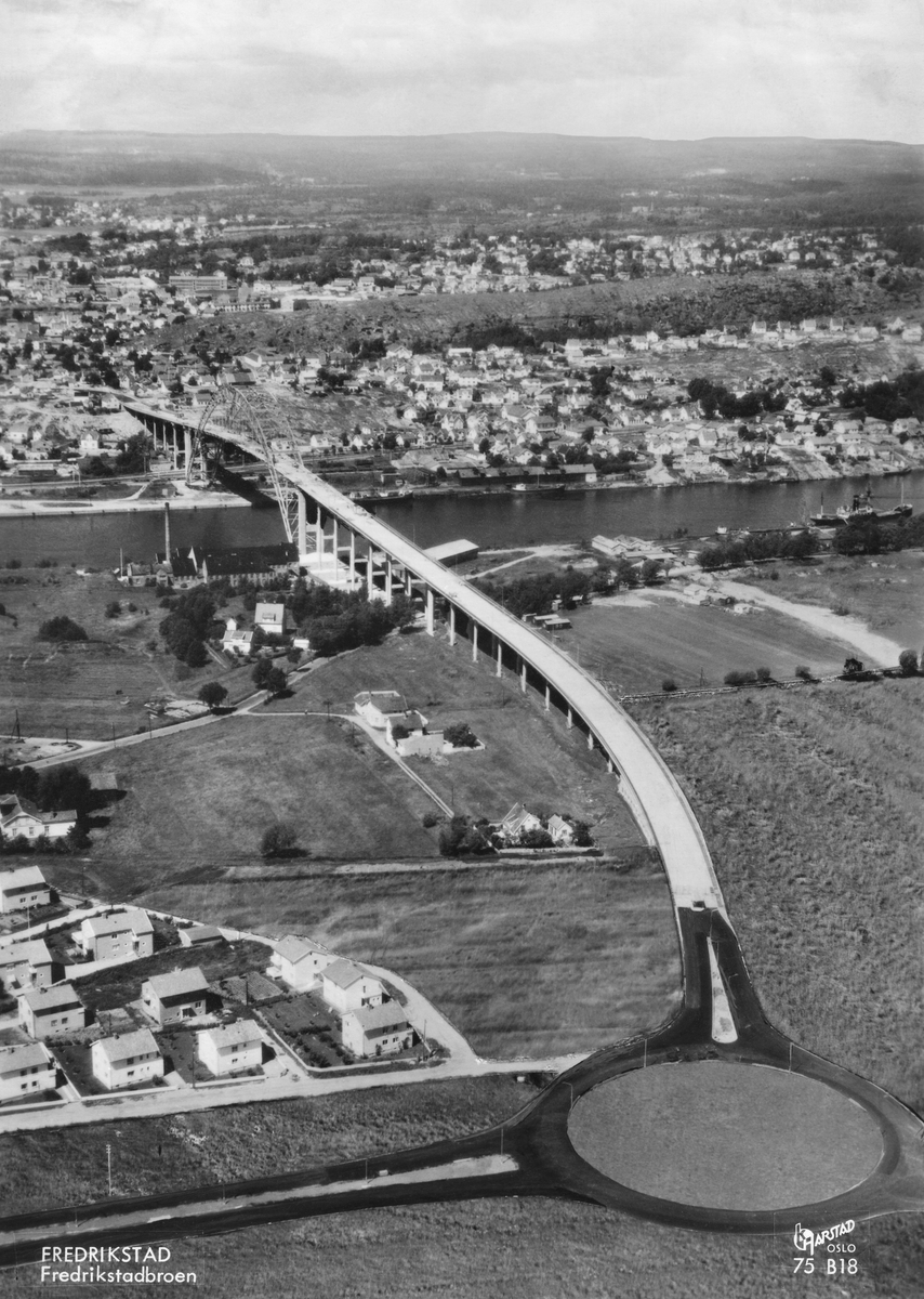 Fredrikstad. Vestsiden lensgt borte, Østsiden nærmest. Fredrikstadbroa. åpnet 18. august 1957. Frem til da hadde all trafikk mellom Vestsiden og Østsiden foregått med ferjer. På Østsiden sees Prestelandet og Gudeberg-området mot elva.  Det norske Cheviotspinneriet til høyre for broa. Broholdet i nedre bildekant var Fredrikstads første rundkjøring og ble tatt i bruk i forbindelse med broåpningen. Nabbetorpveien går under broa, r fra Prestelandet og nordover, frem til tettstedet Nabbetorp. utenfor høyre bildekant. - På Vestsiden ender broa på Kapellfjellet, Mot høyre i bildet Lahellemoen, Lahelle (ved elva) og Kiæråsen.  - Flyfoto: Harstad.