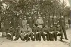 Gruppfoto av en pluton furirer (?) med en löjtnant och en kapten i främre ledet. I bakgrunden syns en nyfiken kvinna i kappa och hatt.