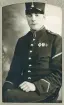En ung man i uniform (m/95, vicekorpral) med kepi och förtjänstmärken på bröstet, poserar hos fotografen.
Knäbild, halvprofil. Ateljéfoto.