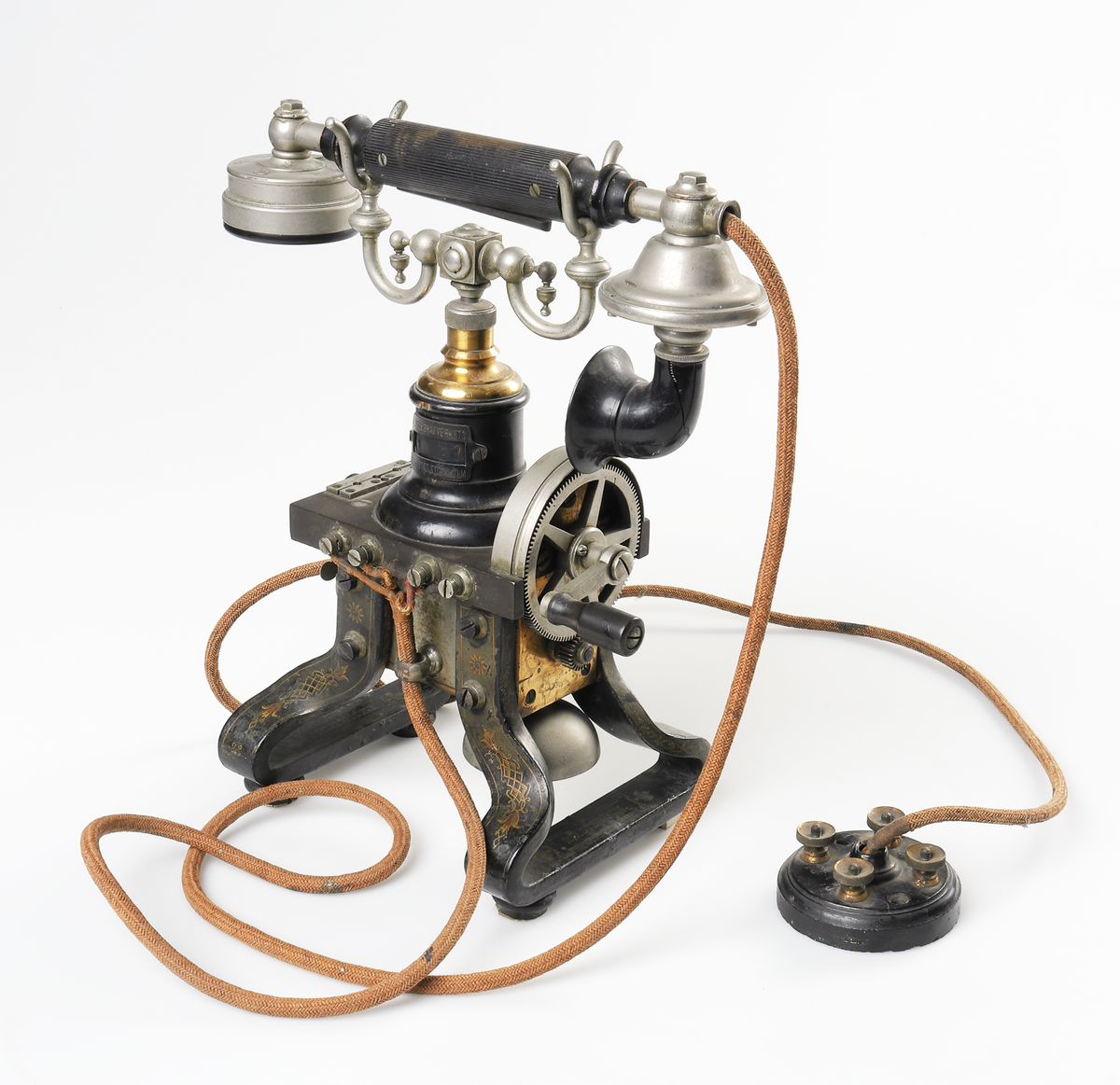 Bordstelefon av Televerkets modell 1892. Bestående av en svartmålad, gulddekorerad ram av plattjärn. Samt däruti infästad en signalgenerator med förnicklad vev och svart knopp. Luren har en svart böjd taltratt samt en samtalsbrytare/sekretessanordning utformad som en svart tangent. Klykan som luren hänger i är förnicklad. Hela telefonen vilar på fyra svarvade tassar av trä. Kabeln mellan luren och spolarna är ljusbrun och tygspunnen.