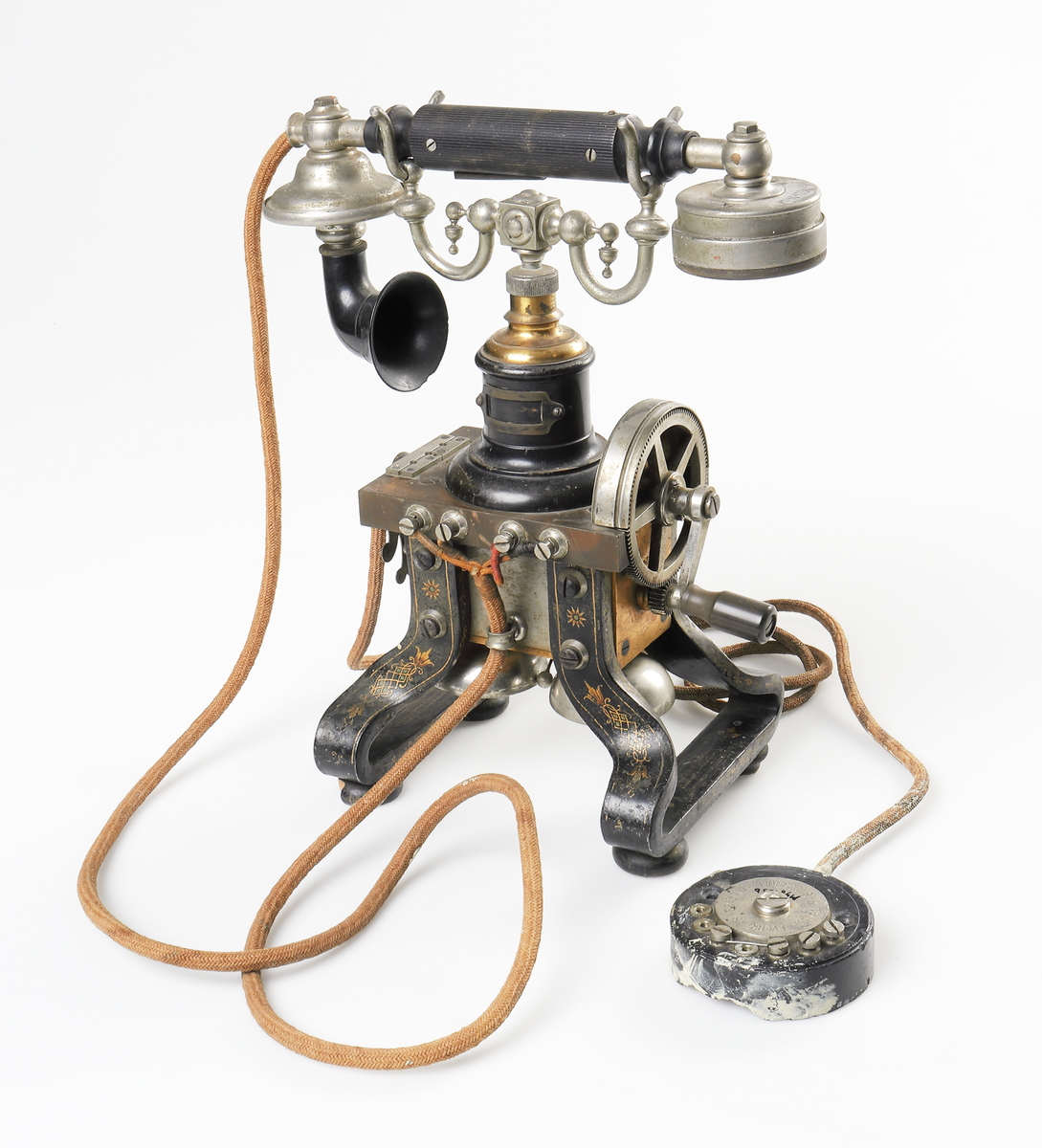Bordstelefon av Televerkets modell 1892. Bestående av en svartmålad, gulddekorerad ram av plattjärn. Samt däruti infästad en signalgenerator med förnicklad vev och svart knopp. Luren har en svart böjd taltratt samt en samtalsbrytare/sekretessanordning utformad som en svart tangent. Klykan som luren hänger i är förnicklad. Hela telefonen vilar på fyra svarvade tassar av trä. Kabeln mellan luren och spolarna är ljusbrun och tygspunnen.