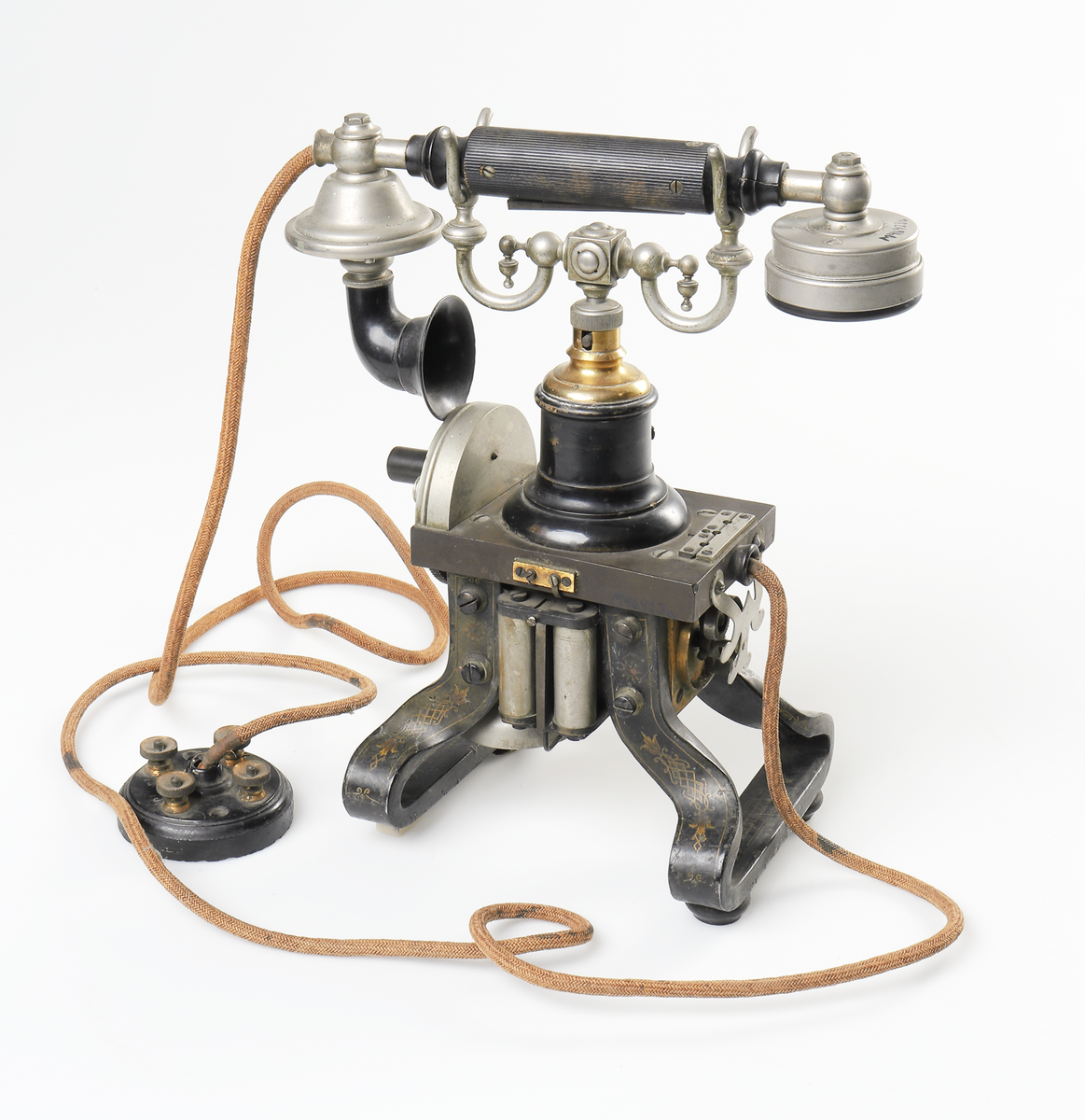 Bordstelefon av Televerkets modell 1892. Bestående av en svartmålad, gulddekorerad ram av plattjärn. Samt däruti infästad en signalgenerator med förnicklad vev och svart knopp. Luren har en svart böjd taltratt samt en samtalsbrytare/sekretessanordning utformad som en svart tangent. Klykan som luren hänger i är förnicklad. Hela telefonen vilar på fyra svarvade tassar av trä. Kabeln mellan luren och spolarna är ljusbrun och tygspunnen.