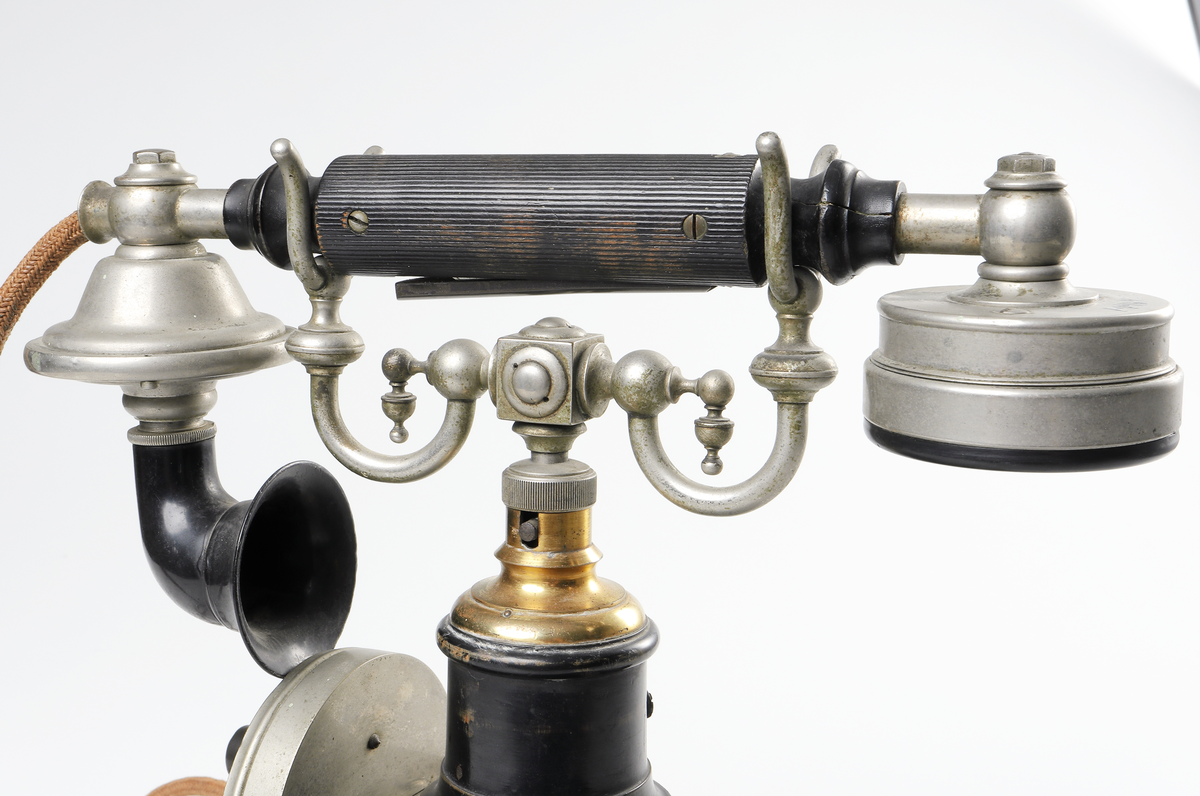 Bordstelefon av Televerkets modell 1892. Bestående av en svartmålad, gulddekorerad ram av plattjärn. Samt däruti infästad en signalgenerator med förnicklad vev och svart knopp. Luren har en svart böjd taltratt samt en samtalsbrytare/sekretessanordning utformad som en svart tangent. Klykan som luren hänger i är förnicklad. Hela telefonen vilar på fyra svarvade tassar av trä. Kabeln mellan luren och spolarna är ljusbrun och tygspunnen.