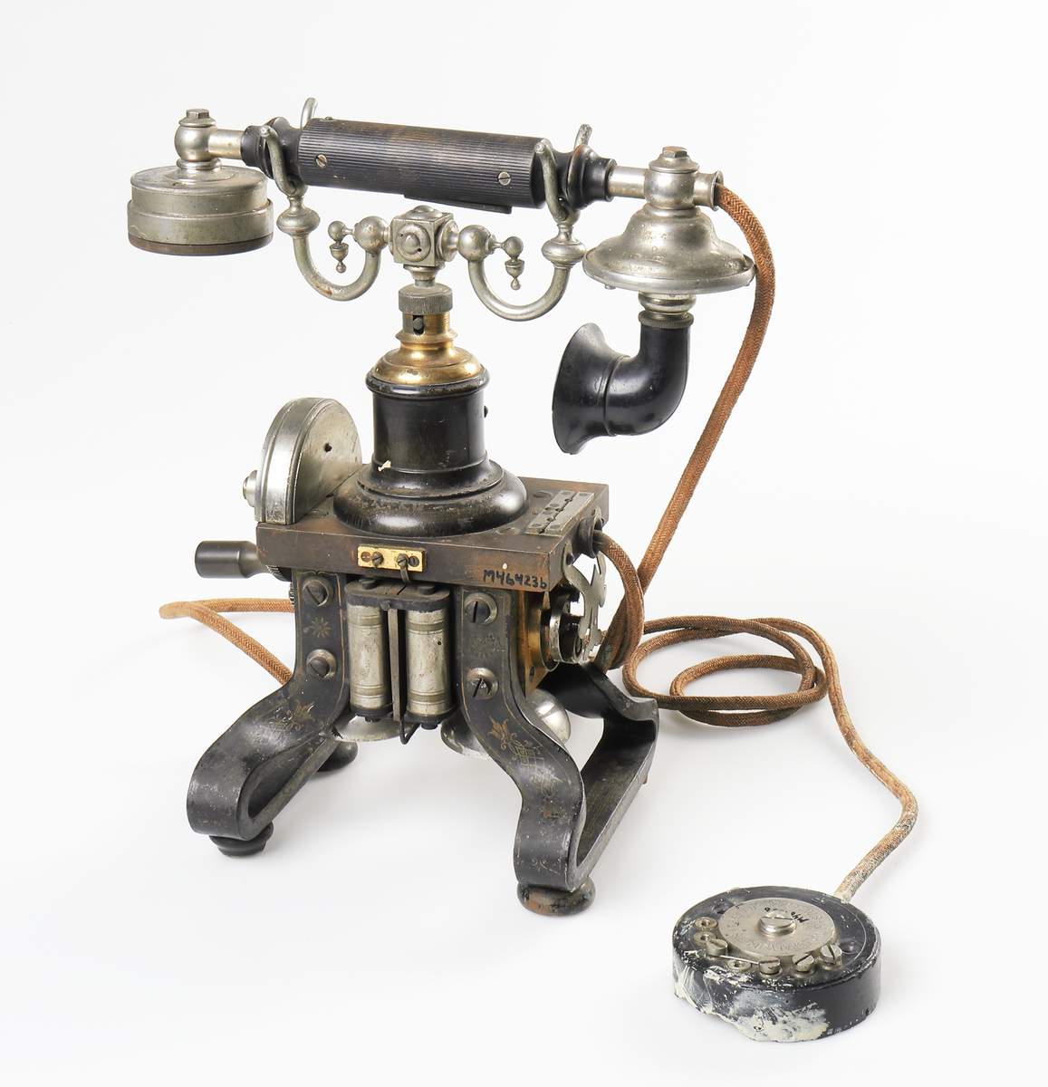 Bordstelefon av Televerkets modell 1892. Bestående av en svartmålad, gulddekorerad ram av plattjärn. Samt däruti infästad en signalgenerator med förnicklad vev och svart knopp. Luren har en svart böjd taltratt samt en samtalsbrytare/sekretessanordning utformad som en svart tangent. Klykan som luren hänger i är förnicklad. Hela telefonen vilar på fyra svarvade tassar av trä. Kabeln mellan luren och spolarna är ljusbrun och tygspunnen.