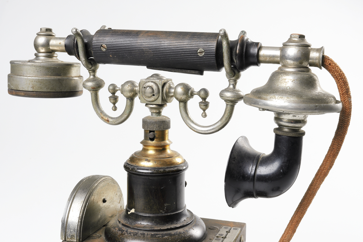 Bordstelefon av Televerkets modell 1892. Bestående av en svartmålad, gulddekorerad ram av plattjärn. Samt däruti infästad en signalgenerator med förnicklad vev och svart knopp. Luren har en svart böjd taltratt samt en samtalsbrytare/sekretessanordning utformad som en svart tangent. Klykan som luren hänger i är förnicklad. Hela telefonen vilar på fyra svarvade tassar av trä. Kabeln mellan luren och spolarna är ljusbrun och tygspunnen.