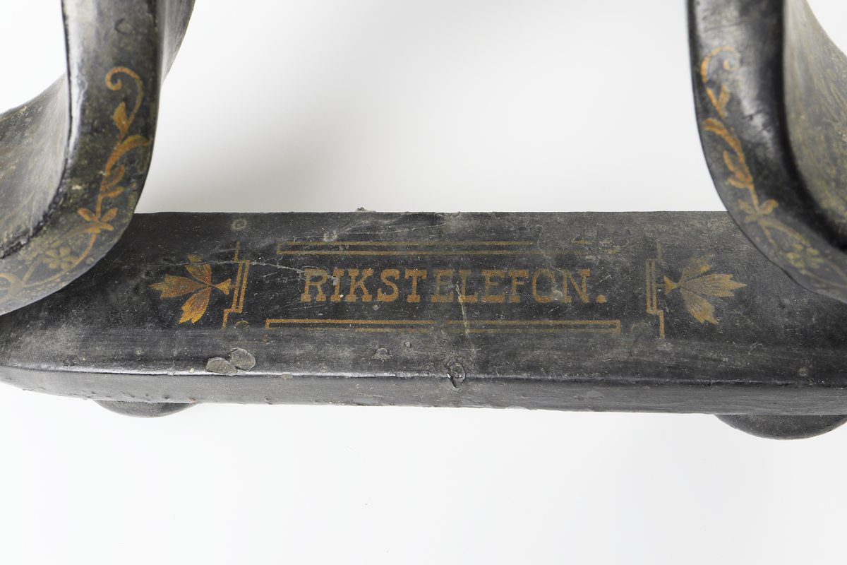 Bordstelefon av Televerkets modell 1892. Bestående av en svartmålad, gulddekorerad ram av plattjärn. Samt däruti infästad en signalgenerator med förnicklad vev och svart knopp. Luren har en svart böjd taltratt samt en samtalsbrytare/sekretessanordning utformad som en svart tangent. Klykan som luren hänger i är förnicklad. Hela telefonen vilar på fyra svarvade tassar av trä. Kabeln mellan luren och spolarna är ljusbrun och tygspunnen.