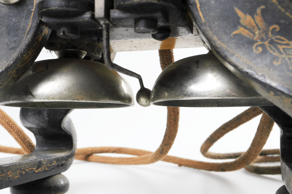 Bordstelefon av Televerkets modell 1892. Bestående av en svartmålad, gulddekorerad ram av plattjärn. Samt däruti infästad en signalgenerator med förnicklad vev och svart knopp. Luren har en svart böjd taltratt samt en samtalsbrytare/sekretessanordning utformad som en svart tangent. Klykan som luren hänger i är förnicklad. Hela telefonen vilar på fyra svarvade tassar av trä. Kabeln mellan luren och spolarna är ljusbrun och tygspunnen.