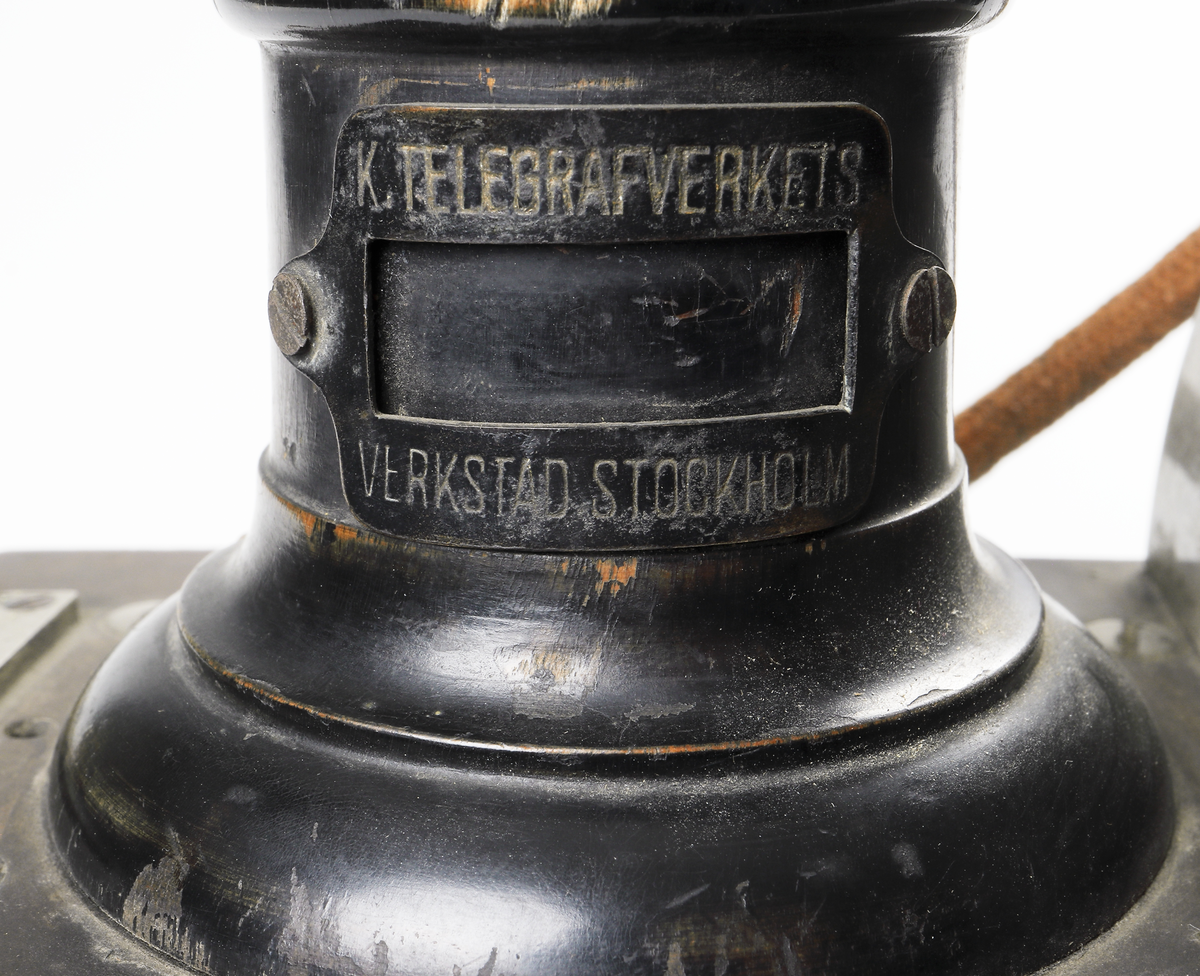 Bordstelefon av Televerkets modell 1892. Bestående av en svartmålad, gulddekorerad ram av plattjärn. Samt däruti infästad en signalgenerator med förnicklad vev och svart knopp. Luren har en svart böjd taltratt samt en samtalsbrytare/sekretessanordning utformad som en svart tangent. Klykan som luren hänger i är förnicklad. Hela telefonen vilar på fyra svarvade tassar av trä. Kabeln mellan luren och spolarna är ljusbrun och tygspunnen.