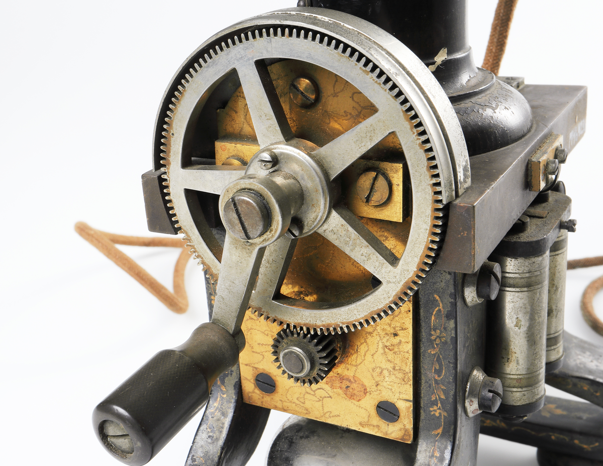 Bordstelefon av Televerkets modell 1892. Bestående av en svartmålad, gulddekorerad ram av plattjärn. Samt däruti infästad en signalgenerator med förnicklad vev och svart knopp. Luren har en svart böjd taltratt samt en samtalsbrytare/sekretessanordning utformad som en svart tangent. Klykan som luren hänger i är förnicklad. Hela telefonen vilar på fyra svarvade tassar av trä. Kabeln mellan luren och spolarna är ljusbrun och tygspunnen.