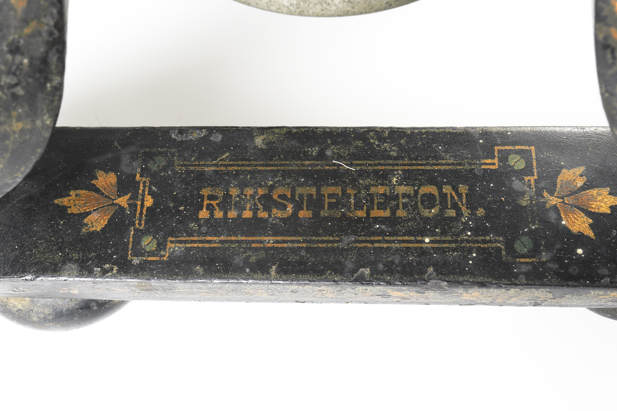 Bordstelefon av Televerkets modell 1892. Bestående av en svartmålad, gulddekorerad ram av plattjärn. Samt däruti infästad en signalgenerator med förnicklad vev och svart knopp. Luren har en svart böjd taltratt samt en samtalsbrytare/sekretessanordning utformad som en svart tangent. Klykan som luren hänger i är förnicklad. Hela telefonen vilar på fyra svarvade tassar av trä. Kabeln mellan luren och spolarna är ljusbrun och tygspunnen.